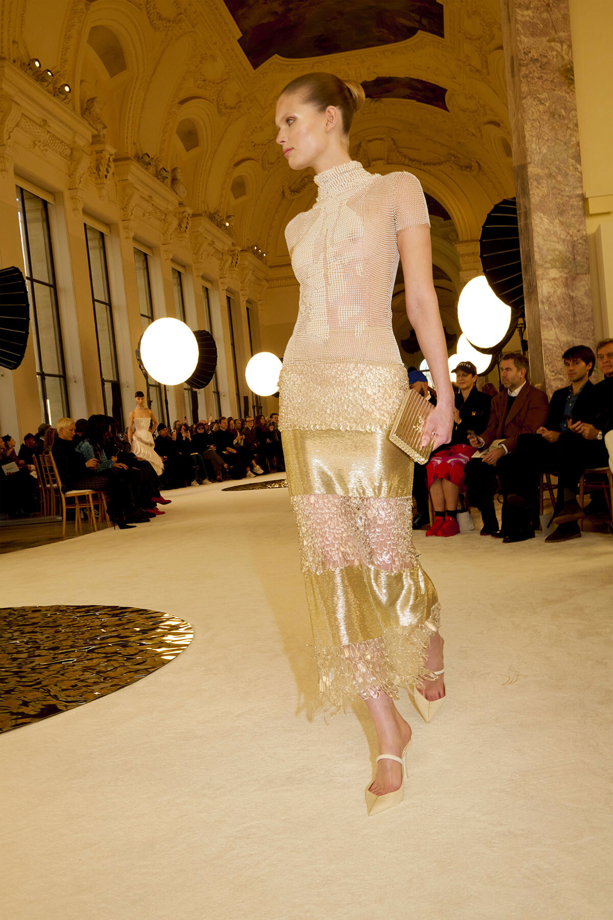Model walking a runway in a pale, beaded top and tiered gold lace skirt inside a grand hall with round stage lights and an audience nearby.