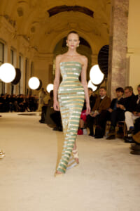 Model walks down a runway wearing a strapless, green-gold striped gown with metallic bands in an ornate hall.