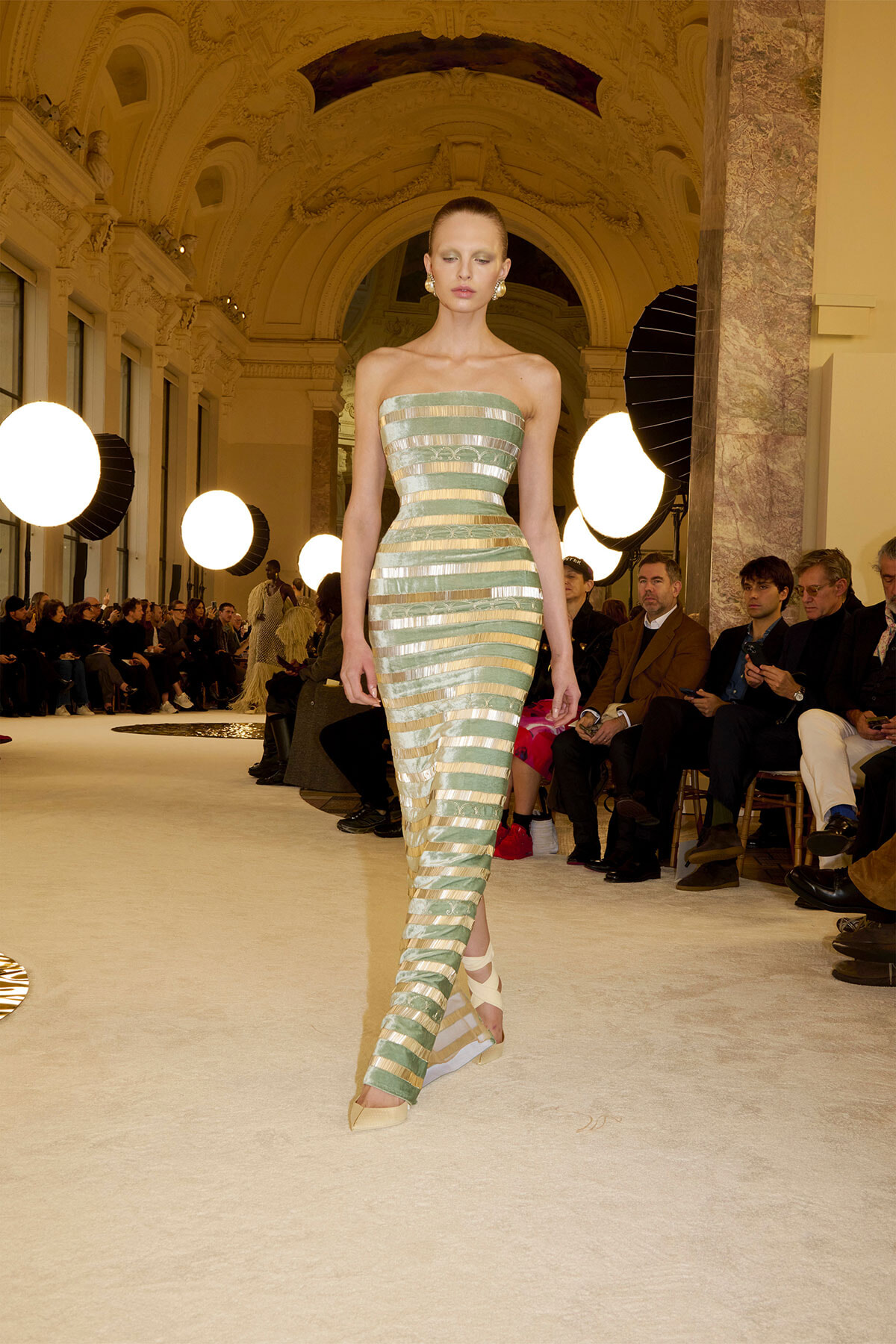 Model walks down a runway wearing a strapless, green-gold striped gown with metallic bands in an ornate hall.