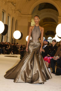 Model walks the runway in a bronze satin gown with a sculpted bodice and sweeping pleated skirt.