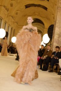 Model wearing a voluminous peach feathered gown on a fashion runway inside an ornate, gilded hall with round studio lights and spectators nearby.