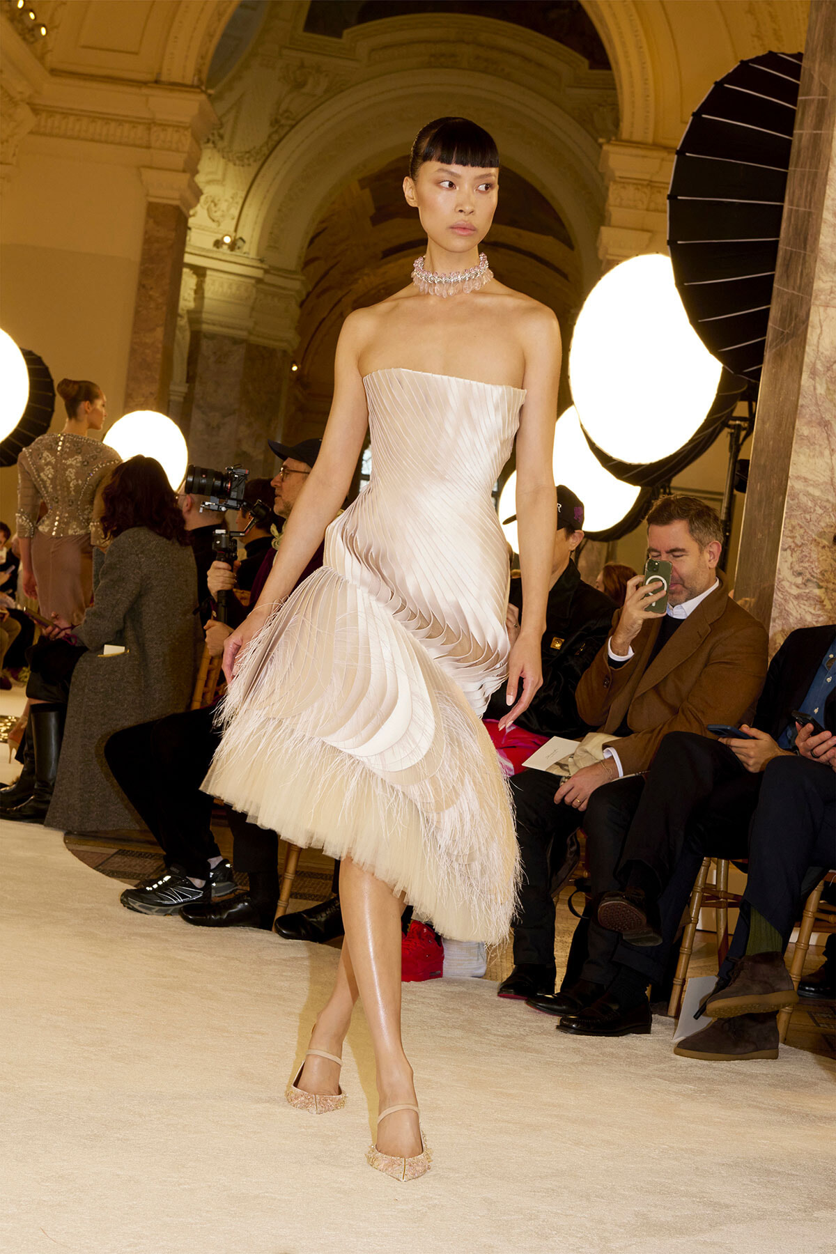 Model in a strapless ivory pleated dress with a layered feathered hem walking a runway at a fashion show, photographers and seated audience in the background.