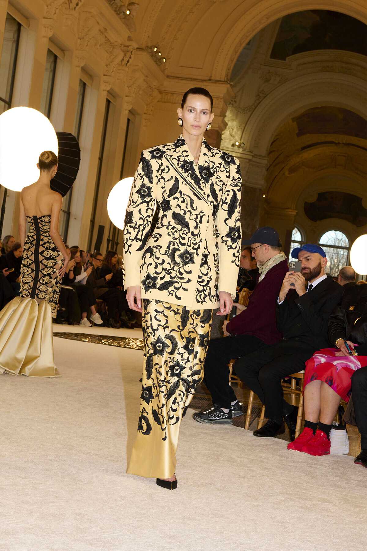 Model on a runway wearing a cream beige suit with black floral embroidery, walking past an audience in a grand, arched hall.