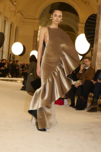 Model walks the runway in a metallic bronze, asymmetrical gown with dramatic drape at a fashion show venue.