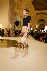 Model walking a runway in a black top and gold embroidered skirt, carrying a small beige handbag in a grand, ornate hall.