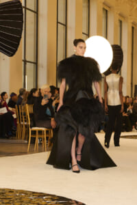 Model in a dramatic black feathered gown walks the runway as audience members and photographers look on in a bright venue.