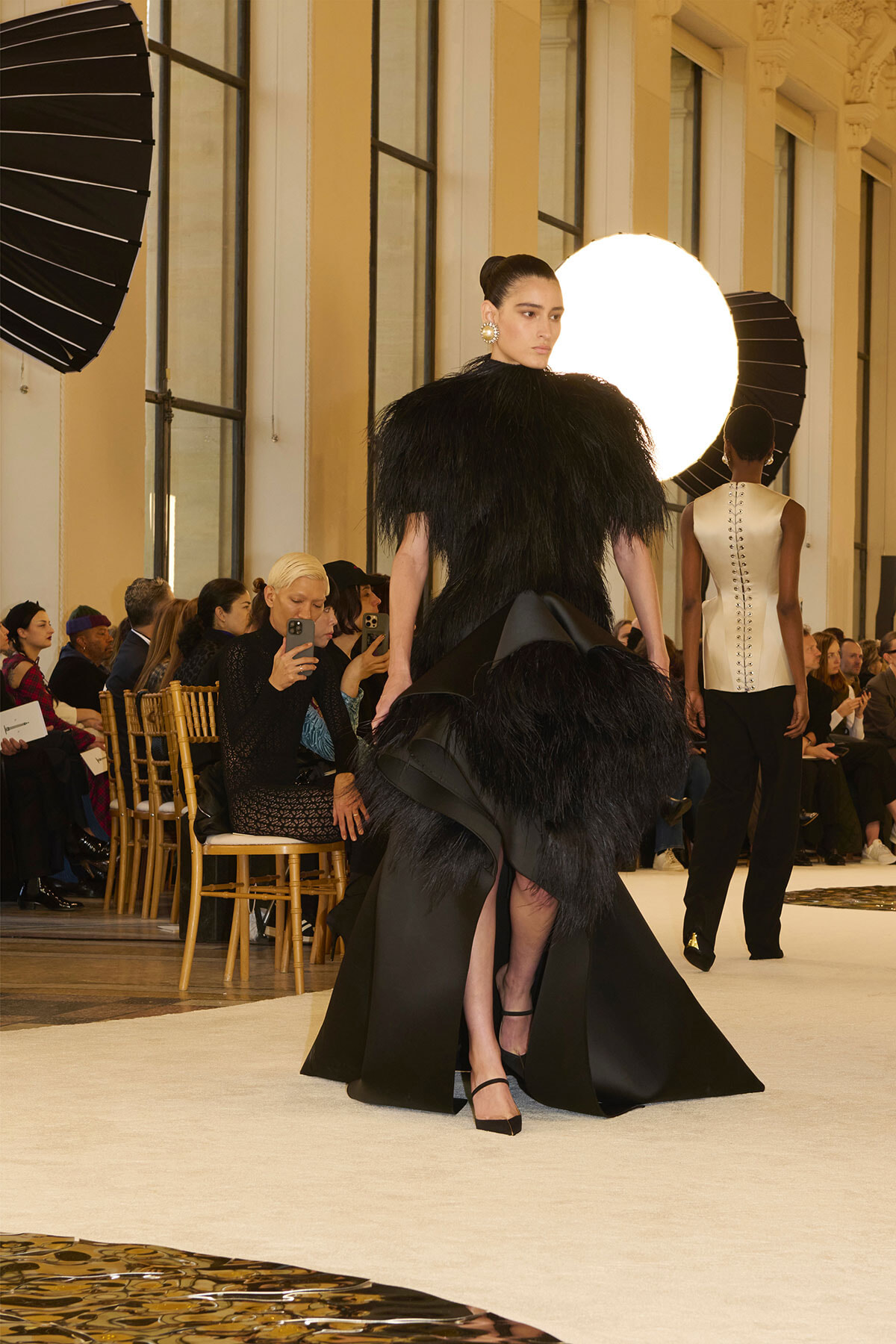 Model in a dramatic black feathered gown walks the runway as audience members and photographers look on in a bright venue.