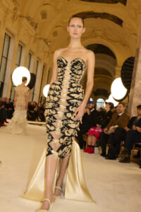 Model walks the runway in a black and gold embroidered strapless gown with sheer panels and a cream satin train in a grand ornate hall