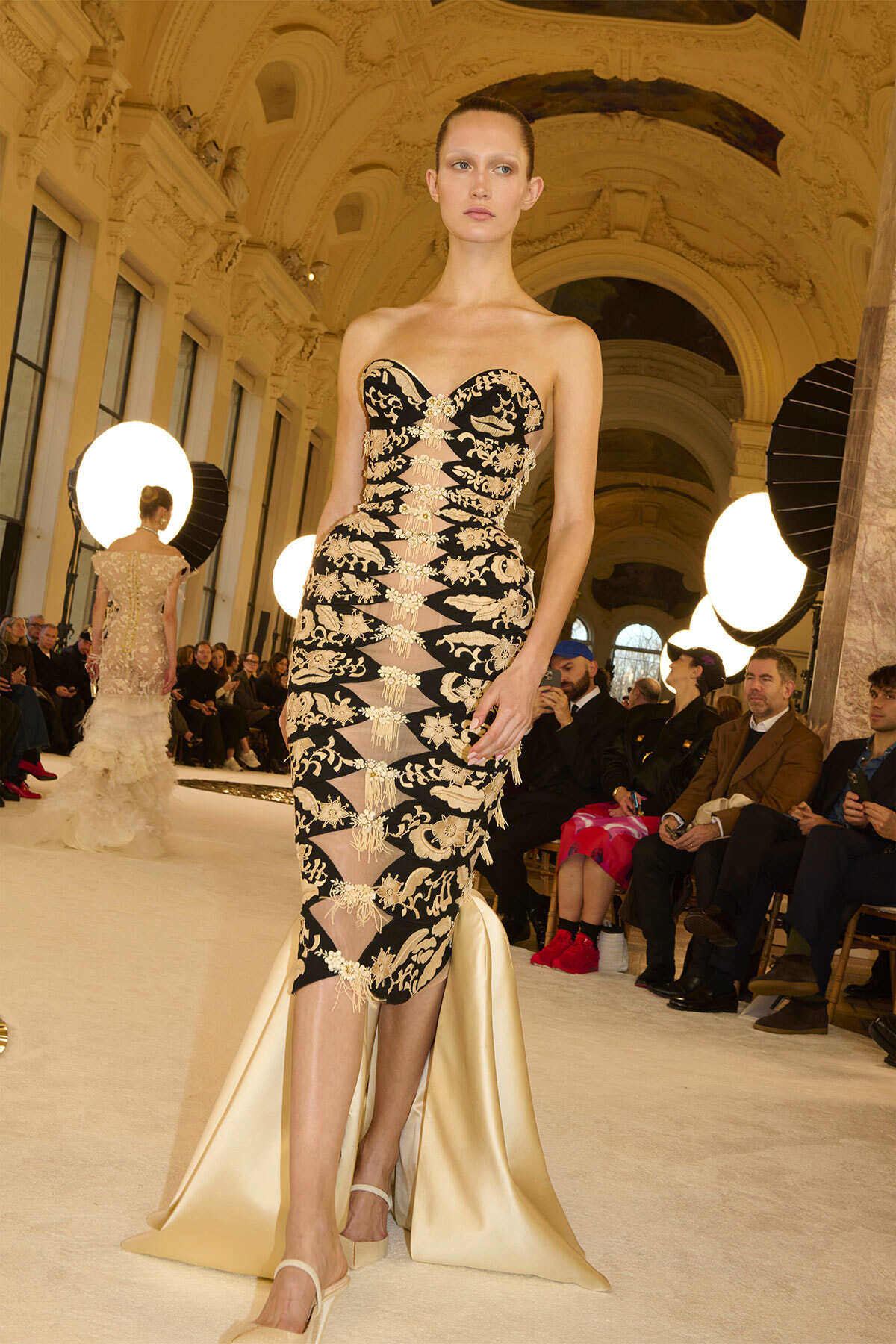 Model walks the runway in a black and gold embroidered strapless gown with sheer panels and a cream satin train in a grand ornate hall