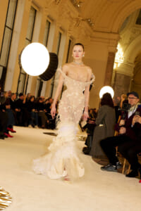 Model walks a runway in an off-shoulder beige lace gown with a feathered train, audience and photographers watching.