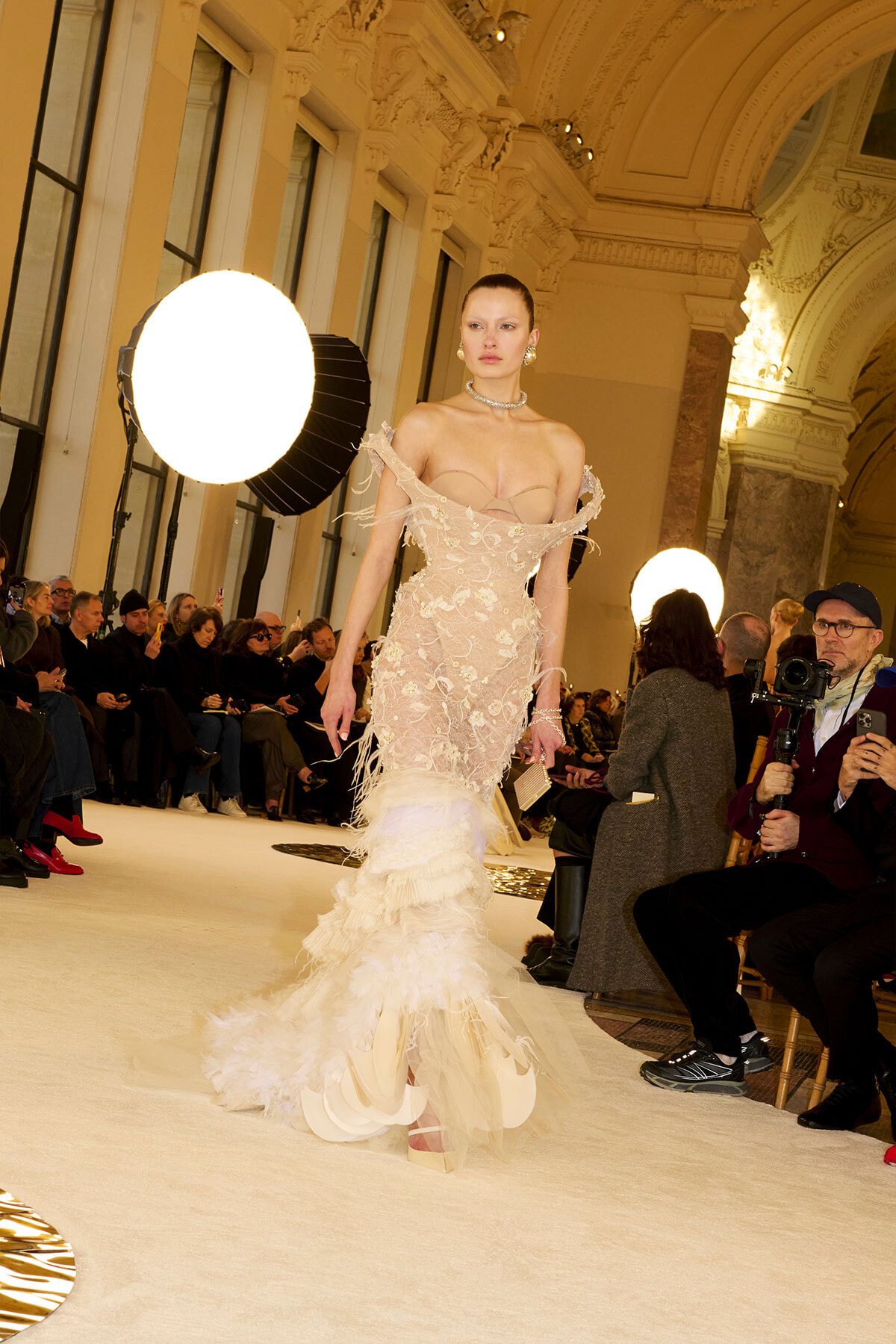 Model walks a runway in an off-shoulder beige lace gown with a feathered train, audience and photographers watching.