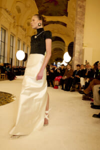 Model walks a runway in a black textured top and white satin skirt, in an ornate hall with spectators on both sides.