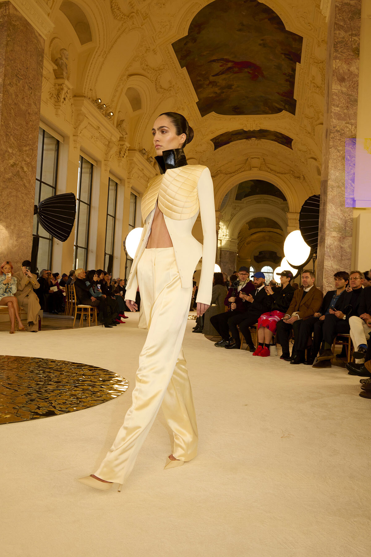 Female model walks a runway in a cream satin suit with quilted shoulder armor and a high black collar, inside an ornate historic hall with an audience lining the runway.