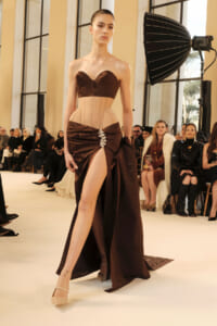 Model on a runway wearing a brown strapless corset and high-slit skirt, posing with a dramatic, structured belt and ornate brooch