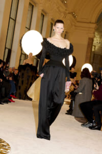 Model walking a fashion runway in a black off-shoulder gown with a beaded ruffled neckline and wide satin pants, in a grand indoor venue with bright circular lights and an audience.