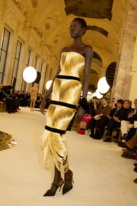 Black model walks a runway in a gold pleated strapless dress with black bands during a fashion show.