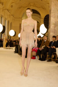 Model walks a beaded strapless mini dress covered in pearls on a runway inside a grand, ornamented hall with bright circular lights and seated audience nearby.