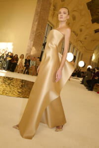 Model walks the runway in a gold strapless satin gown with a dramatic train inside an ornate venue, audience seated along the sides.