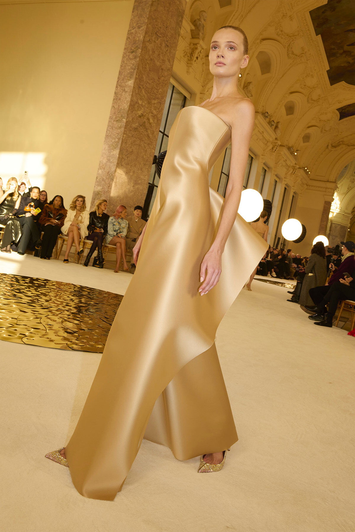 Model walks the runway in a gold strapless satin gown with a dramatic train inside an ornate venue, audience seated along the sides.