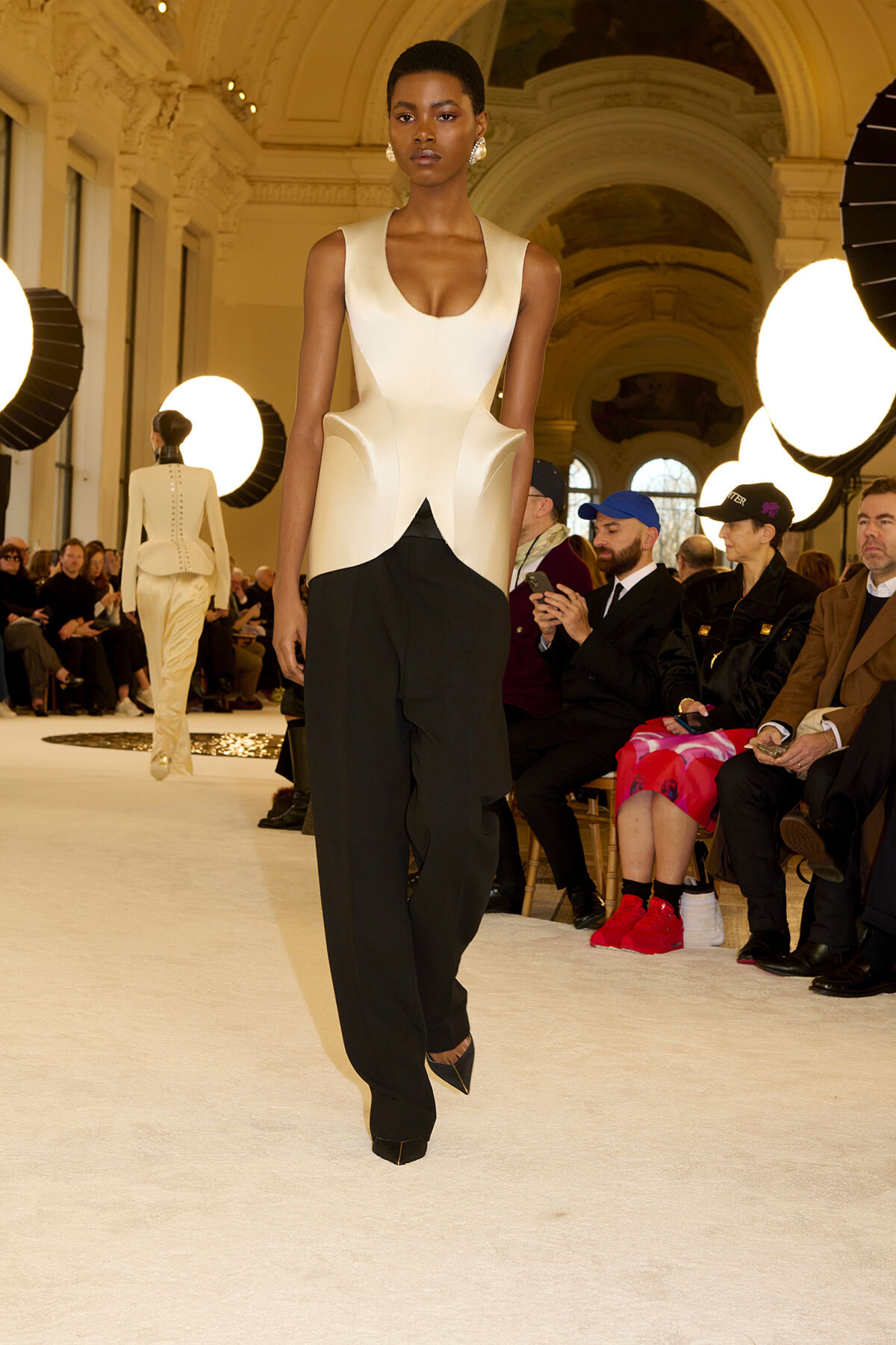 Model walks a runway in a cream sculpted sleeveless top and black wide-leg pants, under bright studio lights in a grand venue.