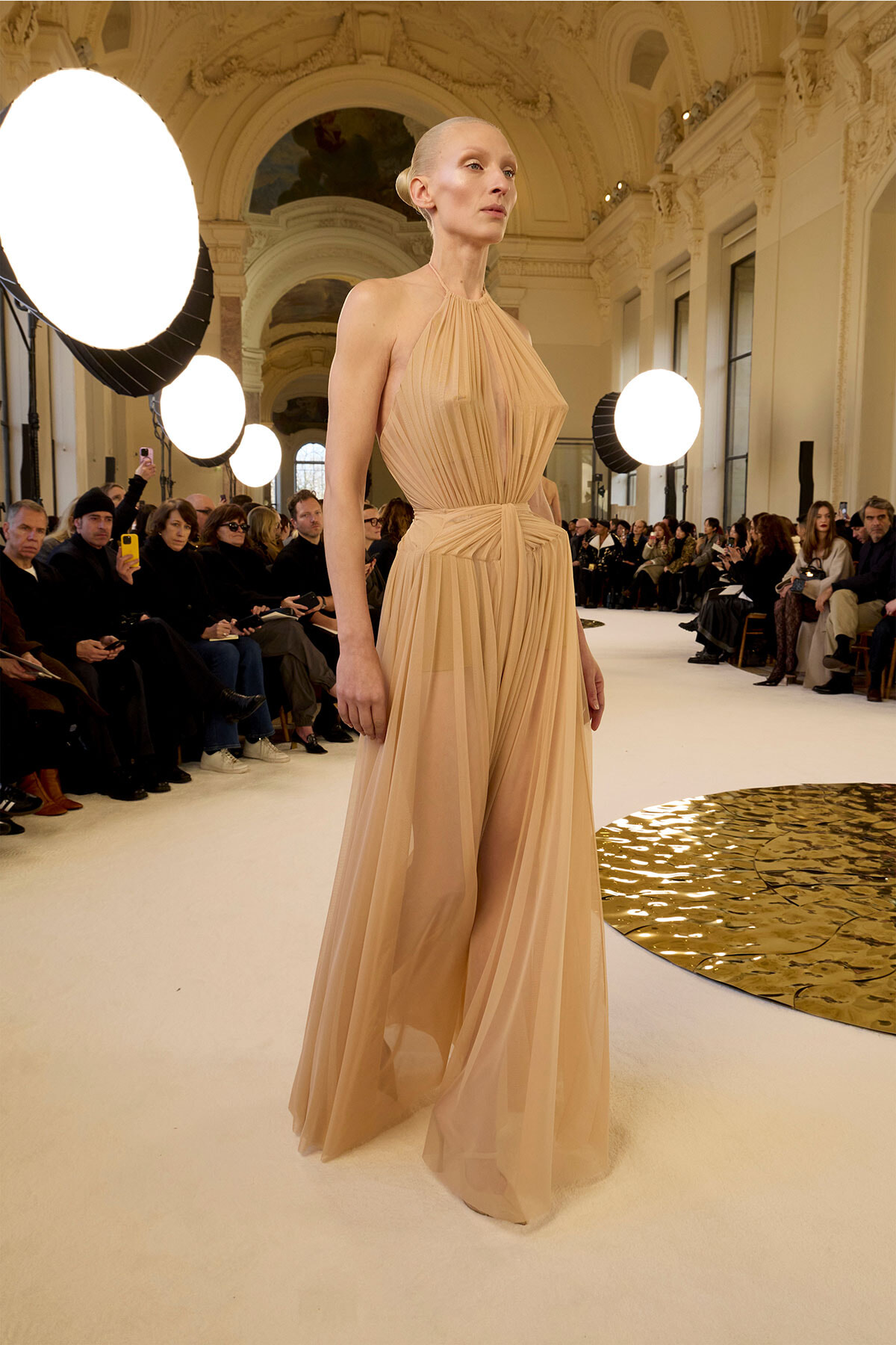 Model walks a nude-toned, pleated halter gown down a fashion-show runway in an ornate hall with seated audience on both sides.]','Beige flowy gown with a high neckline and belt, on a model strutting the catwalk amid spectators.','Elegant nude dress with pleats and a belt, worn by a model at a couture runway show in a grand venue.','Model in a pale peach gown strides the runway; dramatic lighting and ornate interior in the background.','Beige chiffon gown with leg slit and cinched waist on a model at a fashion show, audience watching intently.