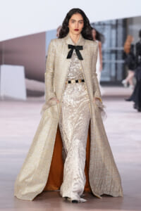 Model walks the runway in a sparkling gold coat over a silver sequin jumpsuit, accented by a black bow at the neck.