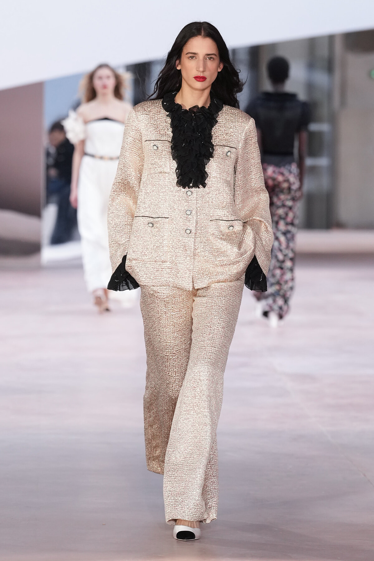 Model walks the runway in a champagne metallic suit with a black ruffled blouse and wide-leg pants, lipstick red.
