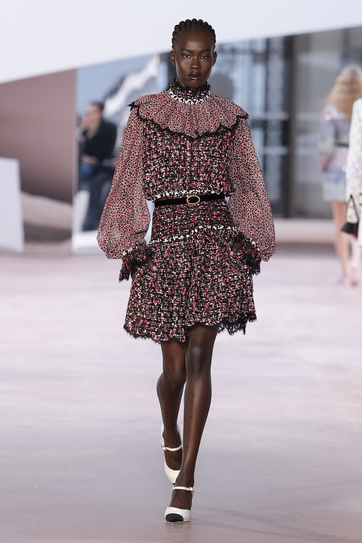 Model walking a runway in a black-pink-white tweed dress with ruffled shoulders, lace trim, and a thin black belt.