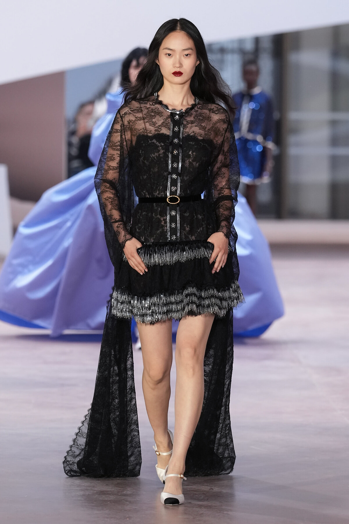 Model walking runway in a black lace dress with sheer long sleeves, gold belt, and layered fringe hem; white heels visible.