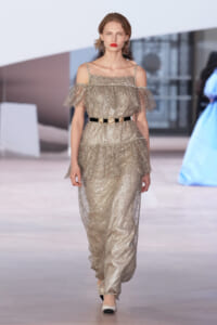 Model wearing a glittery off‑the‑shoulder gown with thin shoulder straps and a black belt featuring gold medallions, walking on a runway.