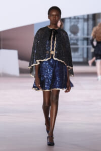 Model on a runway wearing a navy sequined mini dress with a matching cape and gold trim, walking forward confidently.
