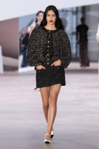 Model walking a fashion runway in a black speckled tweed jacket and matching mini skirt with gold buttons and belt.