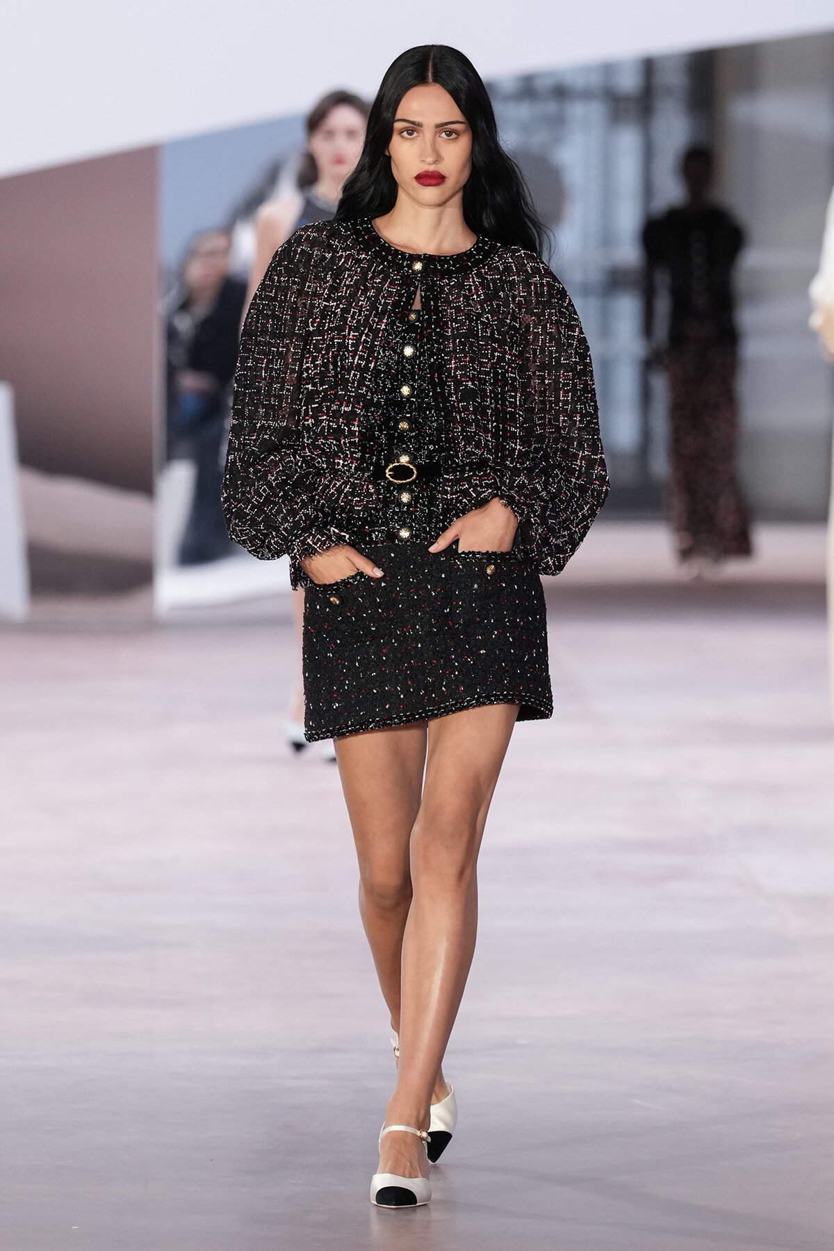 Model walking a fashion runway in a black speckled tweed jacket and matching mini skirt with gold buttons and belt.