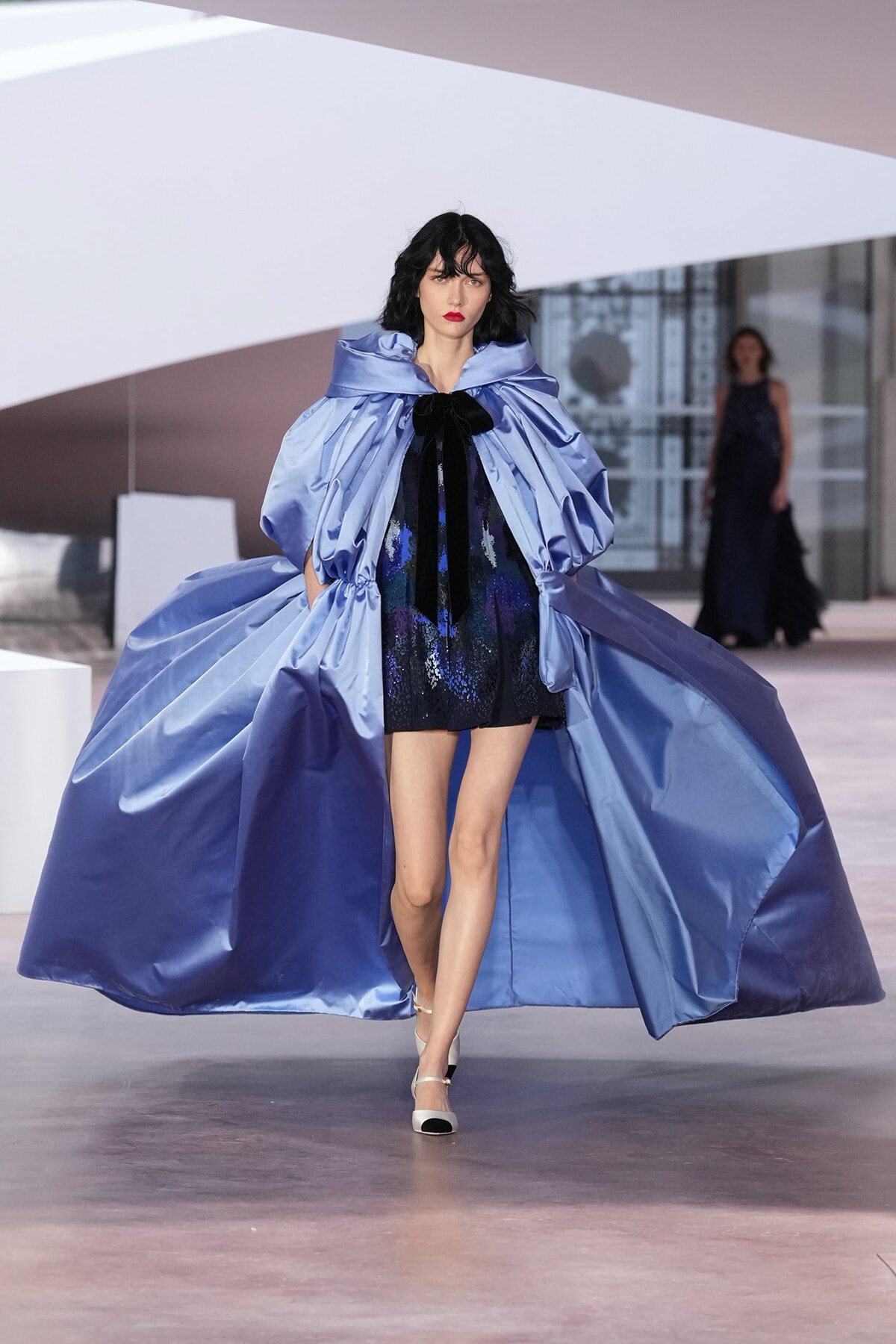 Model walking runway in a glossy light-blue cape over a dark sequined mini dress, posing confidently.