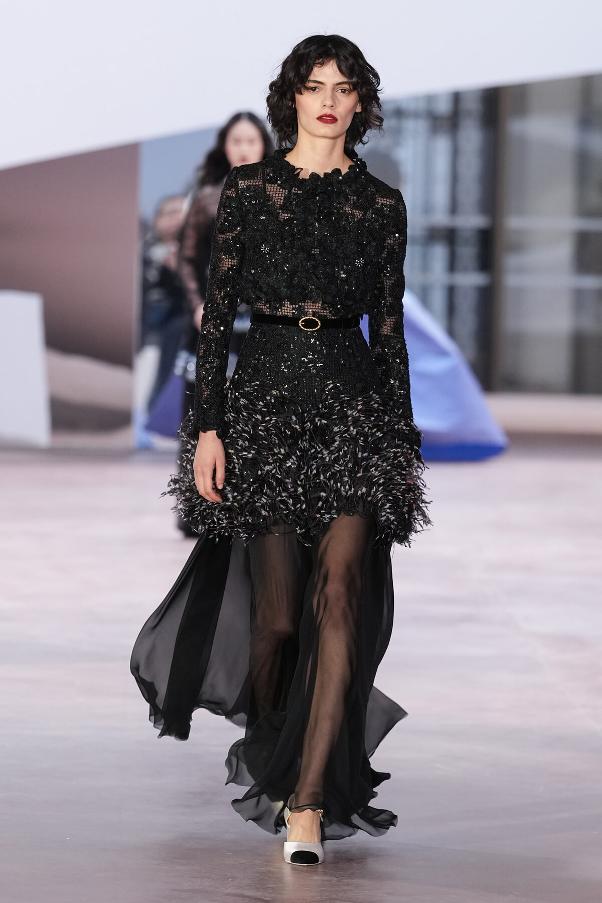 Model walking a runway in a black lace gown with a feathered, voluminous hem and sheer overlay skirt, cinched by a slim belt.