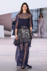 Model walking a runway in a navy sheer blouse over a slip, paired with a glittery, layered skirt and a gold belt.