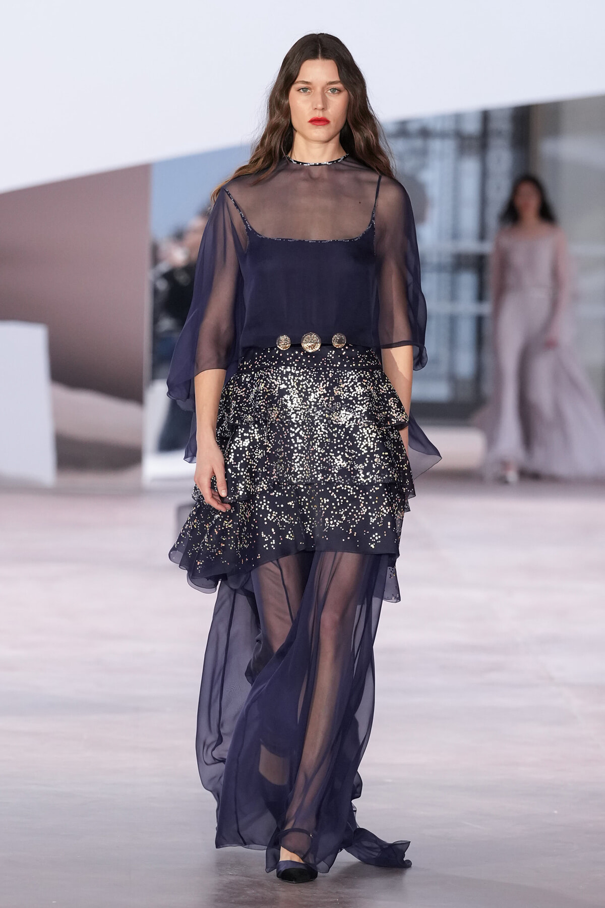 Model walking a runway in a navy sheer blouse over a slip, paired with a glittery, layered skirt and a gold belt.