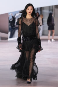 Model walks the runway in a sheer black lace gown with puffed sleeves and a beaded fringe belt at the waist.