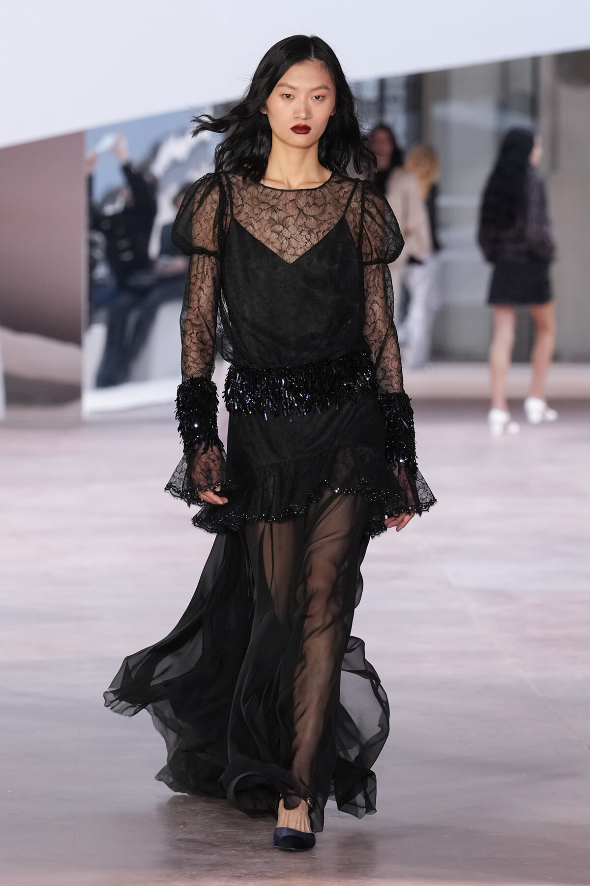 Model walks the runway in a sheer black lace gown with puffed sleeves and a beaded fringe belt at the waist.