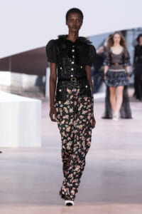 Model on a runway wearing a black sequined blouse with puff sleeves and a black belt, paired with floral-print wide-leg pants.