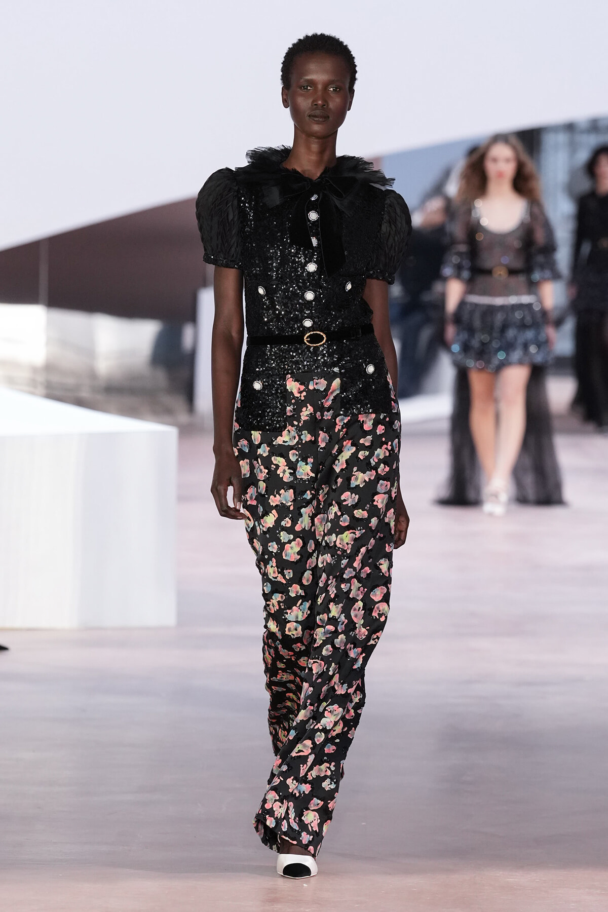 Model on a runway wearing a black sequined blouse with puff sleeves and a black belt, paired with floral-print wide-leg pants.