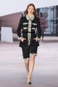 Model walking the runway in a black sequined suit with white floral trim and belt, hands in pockets.