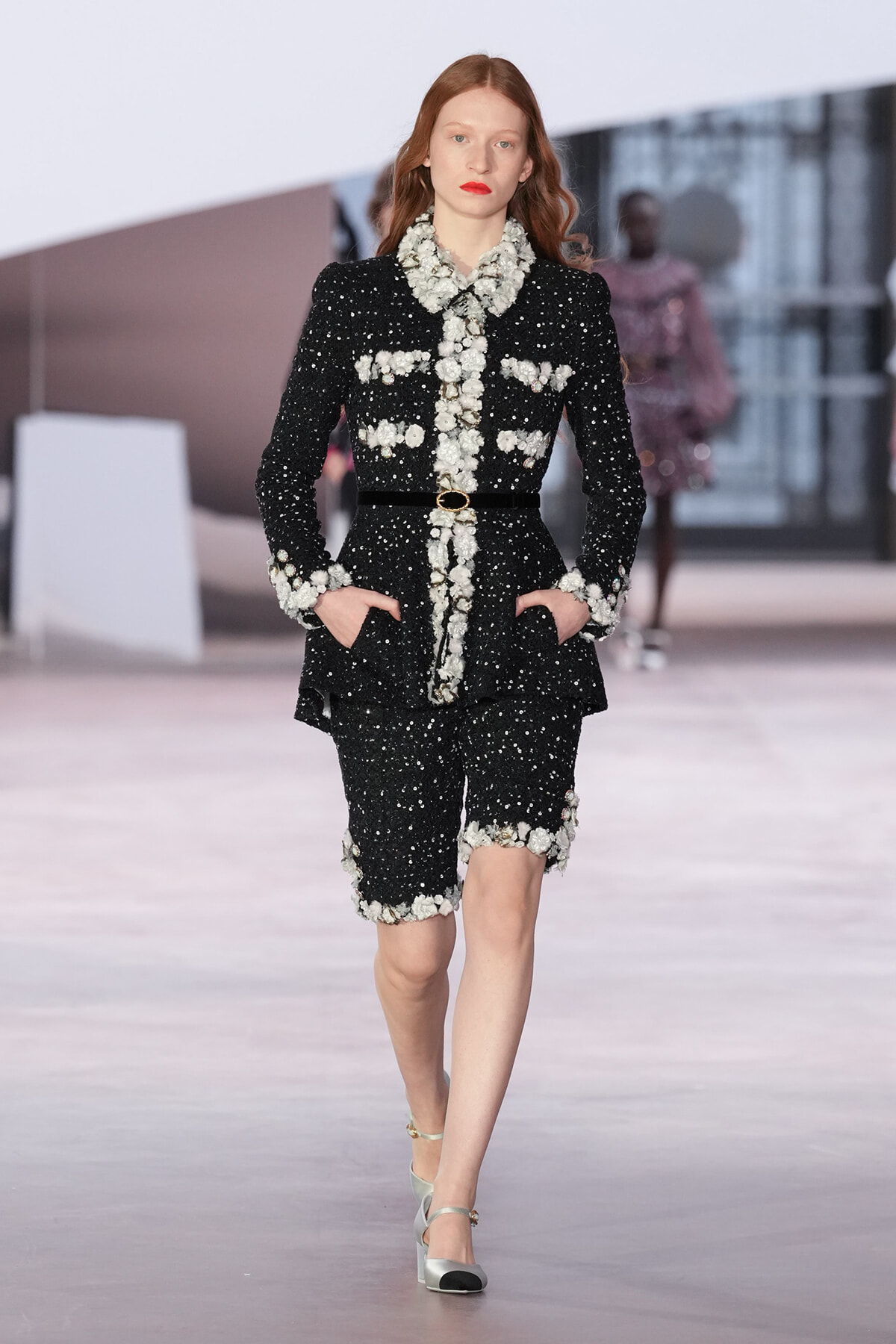 Model walking the runway in a black sequined suit with white floral trim and belt, hands in pockets.