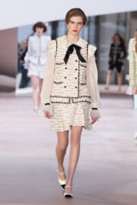 Model walks a fashion runway wearing a cream tweed suit with a black bow at the neck and sheer puff sleeves.
