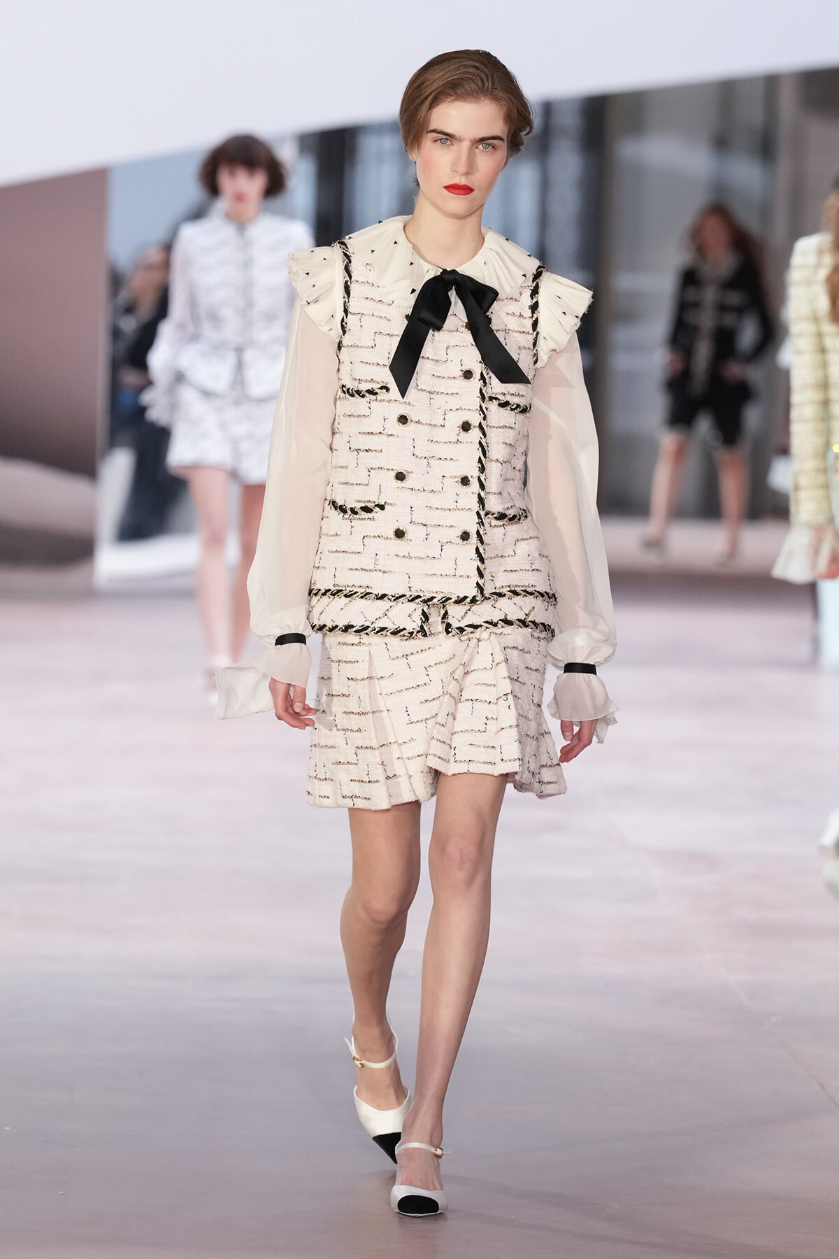 Model walks a fashion runway wearing a cream tweed suit with a black bow at the neck and sheer puff sleeves.