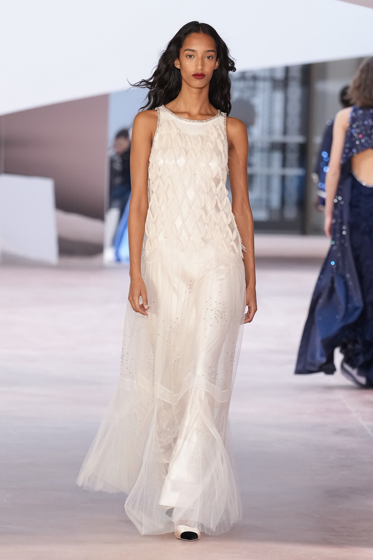 Runway model wearing a sleeveless ivory gown with a diamond-patterned bodice and a sheer, crystal-speckled skirt that flows to the floor.