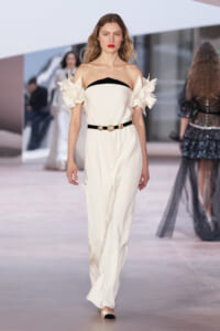 Model on a runway wearing an ivory off-shoulder jumpsuit with a black neckline, cinched waist, and sculptural floral sleeves.