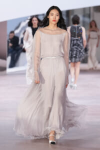Model walks runway in a pale gray, flowing chiffon gown with sheer sleeves and a beaded belt.