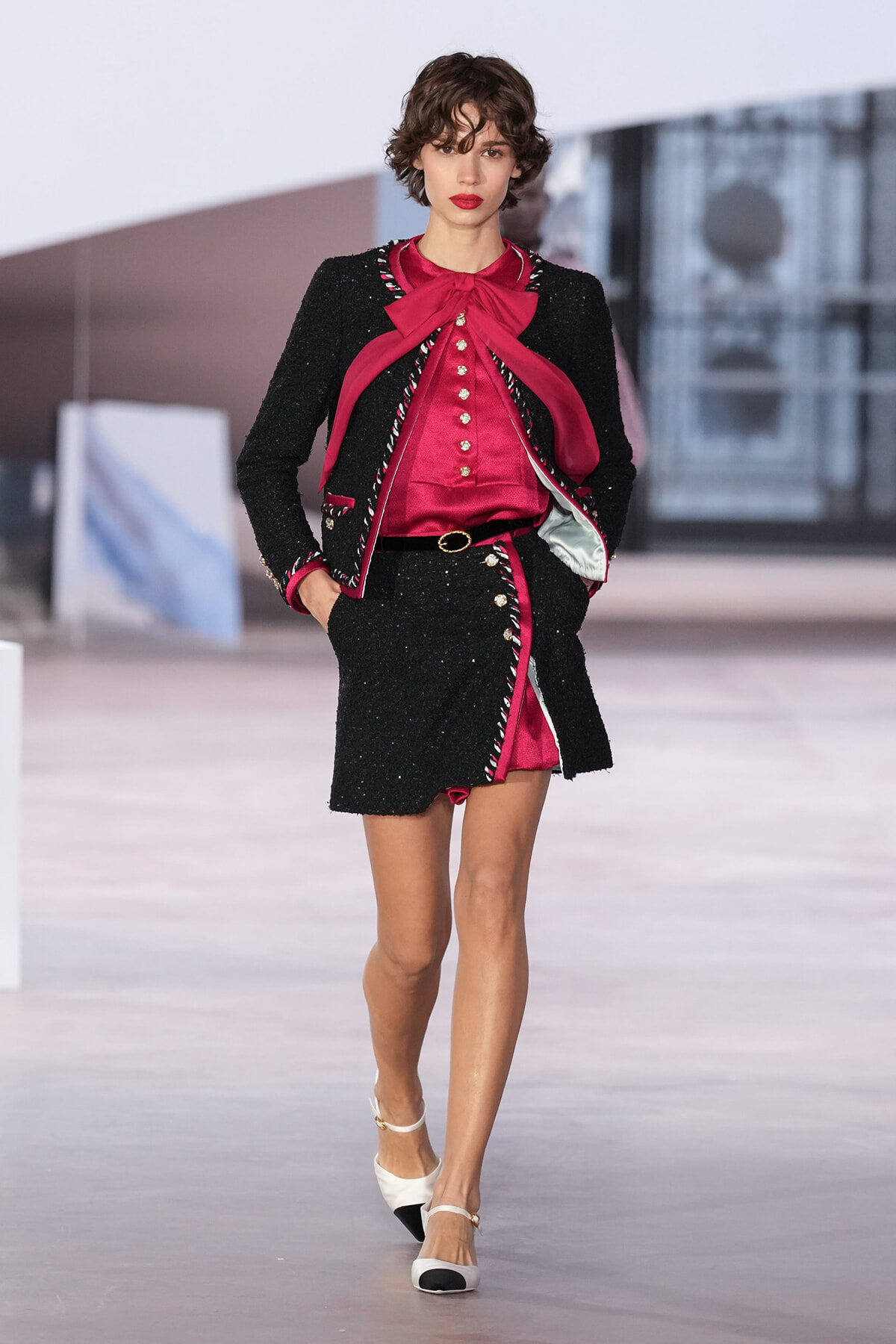 Model walking a runway in a black tweed blazer and matching mini skirt with pink satin blouse featuring a large bow and gold buttons; white-and-black heels complete the look.