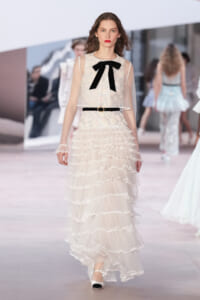 Runway model in a ivory lace gown with sheer sleeves, tiered ruffles, a slim belt, and a black bow at the neckline.